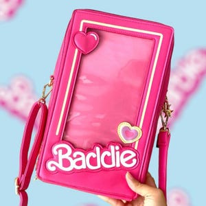 Peut inclure: Sac bandoulière rectangulaire rose vif avec une fenêtre transparente, des cœurs décoratifs et le mot "Baddie" en écriture blanche. Il comprend une bandoulière dorée et une bandoulière rose. Le sac est d'un rose vif.