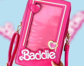 Può includere: Borsa a tracolla rettangolare rosa con finestra trasparente, dettagli a cuore e la scritta "Baddie" in bianco. Include una tracolla a catena dorata e una tracolla rosa. La borsa è di un rosa vibrante.
