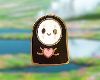 Puede incluir: Un pin de esmalte dorado con un personaje de dibujos animados con una cara en blanco y negro y un corazón rosa. El personaje lleva una túnica negra con detalles dorados.
