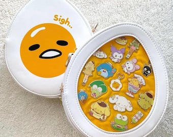 May include: White egg-shaped purse with a cartoon yolk design and the word "Sigh..". The open purse reveals a yellow interior filled with various character pins. The purse has a gold zipper.