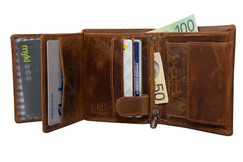 May include: A brown leather wallet with a zipped compartment, multiple card slots, and a flap with a snap closure. The wallet has the text "RFID SAFE" and "Hand Made Full Grain Leather" printed on it. The wallet is open and contains cards and cash.