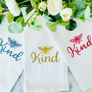 Puede incluir: Tres toallas de cocina blancas con abejas bordadas y la palabra "Kind" en diferentes colores. Las toallas son azules, doradas y rojas.