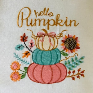 Op de afbeelding: Geborduurd ontwerp op witte stof met gestapelde pompoenen in roze, blauwgroene en crèmetinten, bekroond met een strik. De tekst "hello Pumpkin" is geborduurd in gouden letters, met bloemen- en bladaccenten.