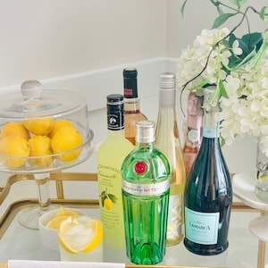 May include: A gold bar cart with a glass top, a glass cloche with lemons, a bottle of limoncello, a bottle of gin, a bottle of white wine, a bottle of sparkling wine, a glass with a lemon wedge, and a glass jar with pink and orange candy.