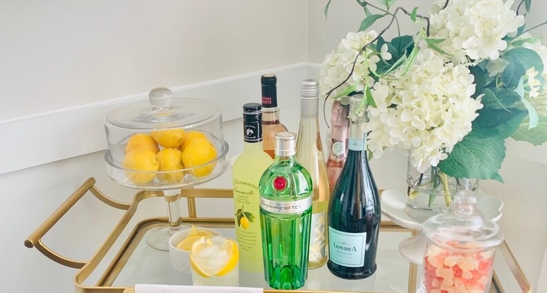 May include: A gold bar cart with a glass top, a glass cloche with lemons, a bottle of limoncello, a bottle of gin, a bottle of white wine, a bottle of sparkling wine, a glass with a lemon wedge, and a glass jar with pink and orange candy.