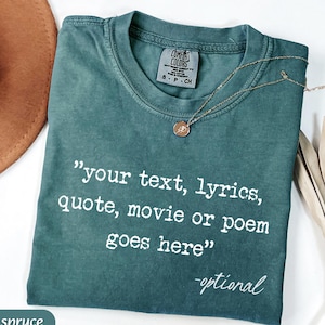 May include: Blue spruce Comfort Colors t-shirt with the text "your text, lyrics, quote, movie or poem goes here" in white. A gold necklace with a pendant is draped on the shirt. The shirt is folded on a white surface.