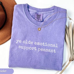May include: A violet-colored t-shirt with the phrase "ye olde emotional support peasant" printed in white. A gold necklace with a pendant rests on the shirt. A brown hat and dried plant are in the background.