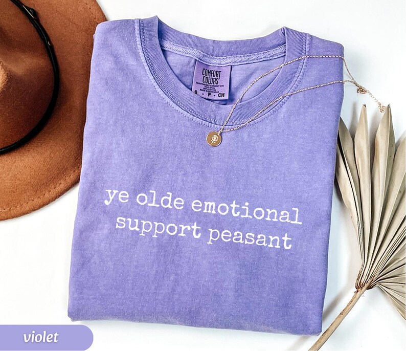 May include: A violet-colored t-shirt with the phrase "ye olde emotional support peasant" printed in white. A gold necklace with a pendant rests on the shirt. A brown hat and dried plant are in the background.