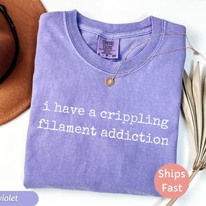 May include: Violet-colored t-shirt with the text "i have a crippling filament addiction" in white. A gold necklace with a pendant is draped on the shirt. The shirt has a Comfort Colors tag.