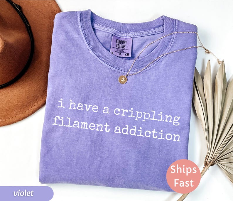 May include: Violet-colored t-shirt with the text "i have a crippling filament addiction" in white. A gold necklace with a pendant is draped on the shirt. The shirt has a Comfort Colors tag.