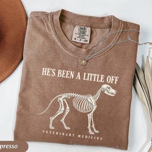 May include: An espresso-colored t-shirt featuring a white graphic of a dog skeleton. The text above the skeleton reads HE'S BEEN A LITTLE OFF and the text below reads VETERINARY MEDICINE. The shirt has a crew neckline and a Comfort Colors brand label.