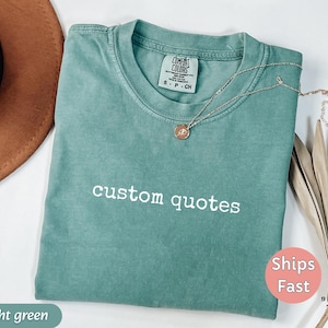 K&ouml;nnte beinhalten: Ein hellgr&uuml;nes T-Shirt mit den Worten "custom quotes" in Wei&szlig; gestickt. Eine silberne Halskette mit einem kleinen Anh&auml;nger liegt auf dem Shirt. Das Shirt ist gefaltet und hat ein Etikett mit der Aufschrift "Comfort Colors".