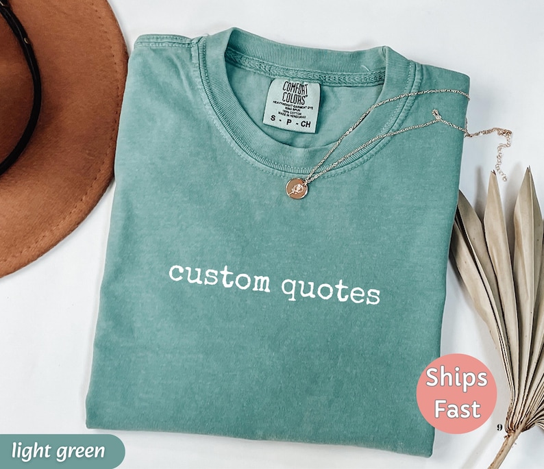 K&ouml;nnte beinhalten: Ein hellgr&uuml;nes T-Shirt mit den Worten "custom quotes" in Wei&szlig; gestickt. Eine silberne Halskette mit einem kleinen Anh&auml;nger liegt auf dem Shirt. Das Shirt ist gefaltet und hat ein Etikett mit der Aufschrift "Comfort Colors".