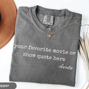 May include: A gray Comfort Colors t-shirt with the text "your favorite movie or show quote here -character" in white. A gold necklace with a pendant is on the shirt. A brown hat is in the background.