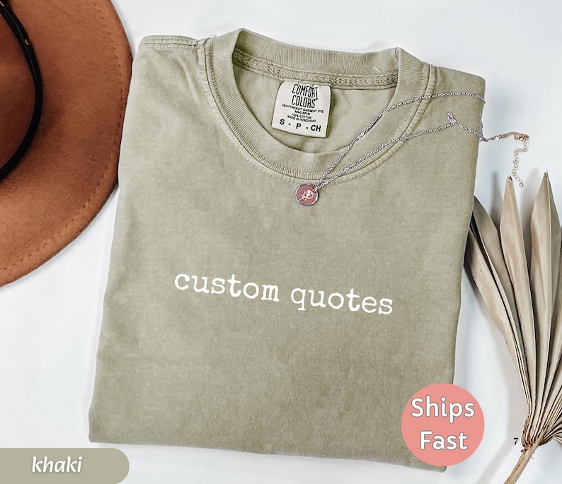K&ouml;nnte beinhalten: Khakifarbenes T-Shirt mit dem wei&szlig;en Text "custom quotes". Das Shirt hat einen Rundhalsausschnitt und kurze &Auml;rmel. Eine silberne Halskette mit einem rosa Anh&auml;nger liegt auf dem Shirt. Ein brauner Hut und eine getrocknete Pflanze sind im Hintergrund.