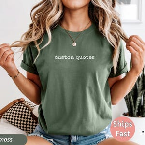 K&ouml;nnte beinhalten: Ein moosgr&uuml;nes T-Shirt mit dem wei&szlig;en Text "custom quotes". Das Shirt hat hochgekrempelte &Auml;rmel und wird mit blauen Jeansshorts getragen. Eine braune und schwarze karierte Tasche ist sichtbar.