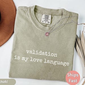 May include: Khaki-colored Comfort Colors t-shirt with the phrase "validation is my love language" printed in white. A silver necklace with a pink pendant rests on the shirt. The shirt is folded and ready to wear.