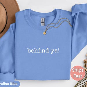 May include: Carolina blue sweatshirt with the text "behind ya!" in white. A gold necklace with a star pendant rests on the sweatshirt. A brown hat is visible in the upper left corner.