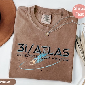 May include: Espresso-colored t-shirt with the text "31/ATLAS INTERSTELLAR VISITOR" in black and teal. The shirt has a crew neck and a small tag. A silver necklace with a pendant is on the shirt.