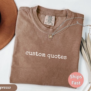K&ouml;nnte beinhalten: Espressofarbenes T-Shirt mit den wei&szlig;en Worten "custom quotes". Das Shirt hat einen Rundhalsausschnitt und ein kleines Etikett am Ausschnitt. Eine silberne Halskette mit Anh&auml;nger liegt auf dem Shirt. Ein brauner Hut und eine getrocknete Pflanze sind im Hintergrund.
