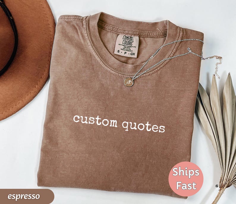 K&ouml;nnte beinhalten: Espressofarbenes T-Shirt mit den wei&szlig;en Worten "custom quotes". Das Shirt hat einen Rundhalsausschnitt und ein kleines Etikett am Ausschnitt. Eine silberne Halskette mit Anh&auml;nger liegt auf dem Shirt. Ein brauner Hut und eine getrocknete Pflanze sind im Hintergrund.