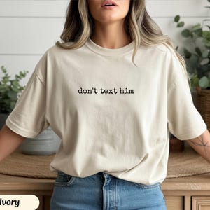 May include: Ivory-colored t-shirt with the text "don't text him" in black. The shirt is a relaxed fit and made of a soft material. The shirt is paired with blue jeans.