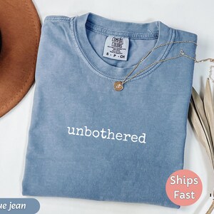 May include: A blue jean colored Comfort Colors t-shirt with the word "unbothered" in white script. A gold necklace with a pendant rests on the shirt. A brown hat is in the upper left corner.