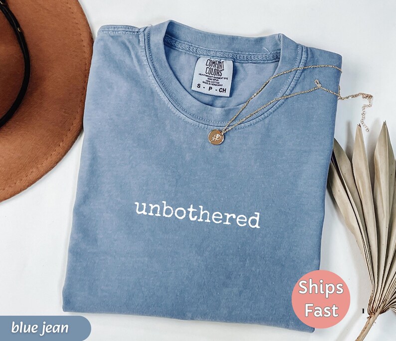 May include: A blue jean colored Comfort Colors t-shirt with the word "unbothered" in white script. A gold necklace with a pendant rests on the shirt. A brown hat is in the upper left corner.
