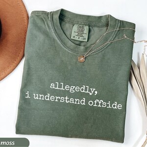 May include: A moss green t-shirt with the text "allegedly, i understand offside" in white. A gold necklace with a pendant rests on the shirt. The shirt is folded on a white surface.