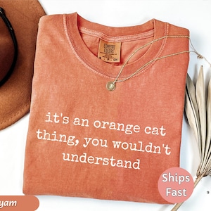 May include: Orange Comfort Colors t-shirt with the text "it's an orange cat thing, you wouldn't understand" in white. A gold necklace with a pendant is on the shirt. A brown hat is visible in the background.