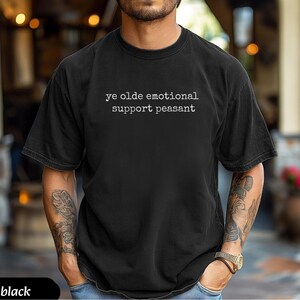 May include: Black t-shirt with the text "ye olde emotional support peasant" in white. The shirt is a dark color and has a relaxed fit. The person wearing the shirt has tattoos on their arms.
