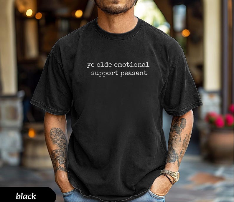 May include: Black t-shirt with the text "ye olde emotional support peasant" in white. The shirt is a dark color and has a relaxed fit. The person wearing the shirt has tattoos on their arms.