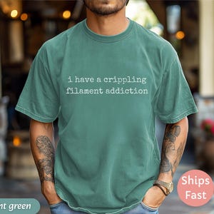 May include: Light green t-shirt with the text "i have a crippling filament addiction" in white. The shirt is a solid color and has short sleeves. The person is wearing a watch and has tattoos on their arms.