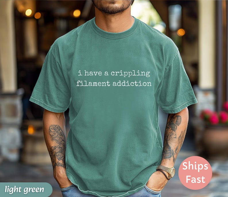 May include: Light green t-shirt with the text "i have a crippling filament addiction" in white. The shirt is a solid color and has short sleeves. The person is wearing a watch and has tattoos on their arms.