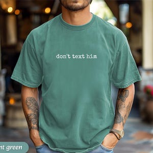 May include: Light green t-shirt with the text "don't text him" in white. The shirt is a solid color and has a relaxed fit. The model is wearing a gold watch and blue jeans.