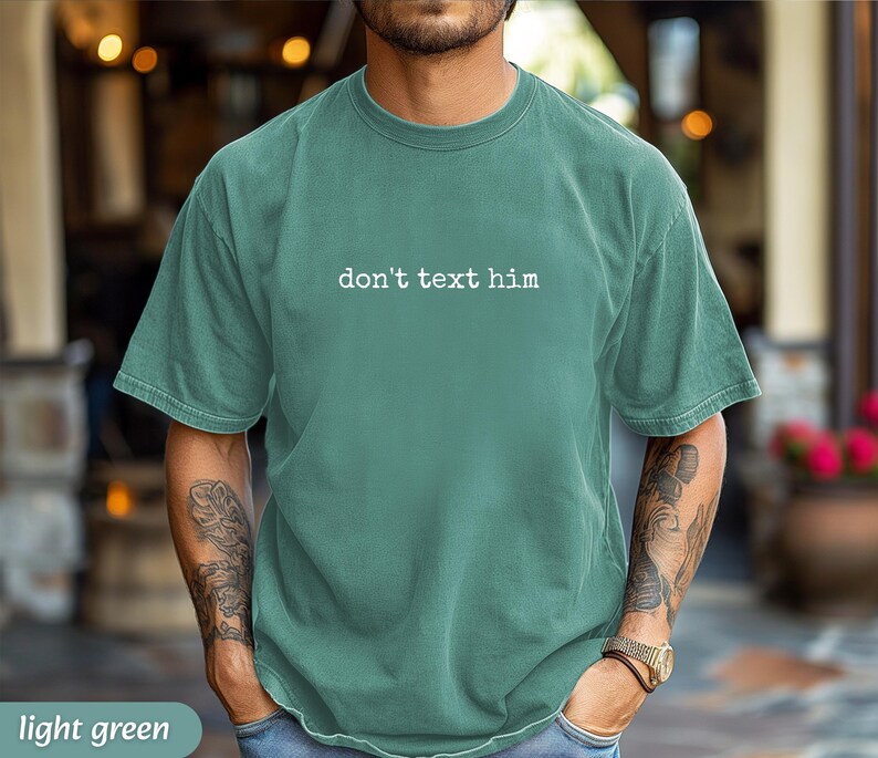 May include: Light green t-shirt with the text "don't text him" in white. The shirt is a solid color and has a relaxed fit. The model is wearing a gold watch and blue jeans.