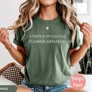 May include: Moss green t-shirt with the text "I have a crippling filament addiction" in white. The shirt has rolled-up sleeves and is worn with blue denim shorts. A small gold necklace is visible.
