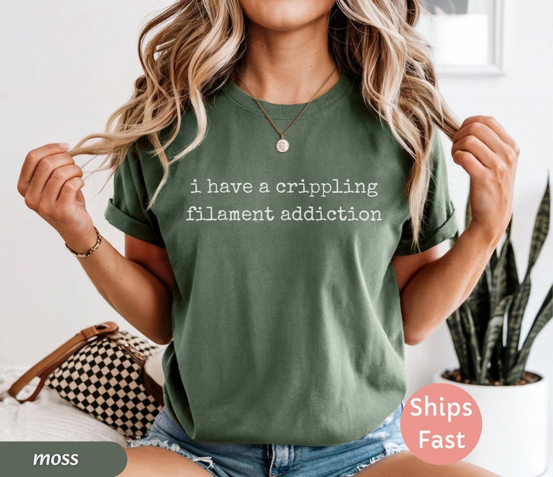 May include: Moss green t-shirt with the text "I have a crippling filament addiction" in white. The shirt has rolled-up sleeves and is worn with blue denim shorts. A small gold necklace is visible.