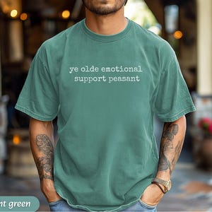 May include: Light green t-shirt with the text "ye olde emotional support peasant" in white. The shirt has short sleeves and a crew neck. The model is wearing a watch and has tattoos on their arms.