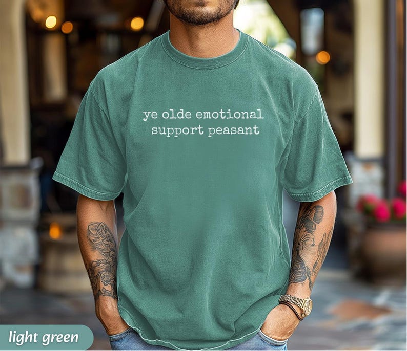 May include: Light green t-shirt with the text "ye olde emotional support peasant" in white. The shirt has short sleeves and a crew neck. The model is wearing a watch and has tattoos on their arms.