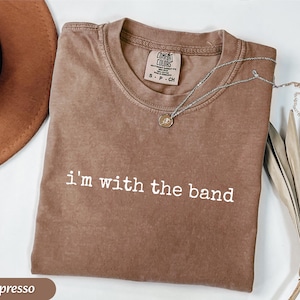 May include: Espresso-colored t-shirt with the text "i'm with the band" in white. The shirt has a crew neck and short sleeves. A silver necklace with a pendant is draped across the shirt.