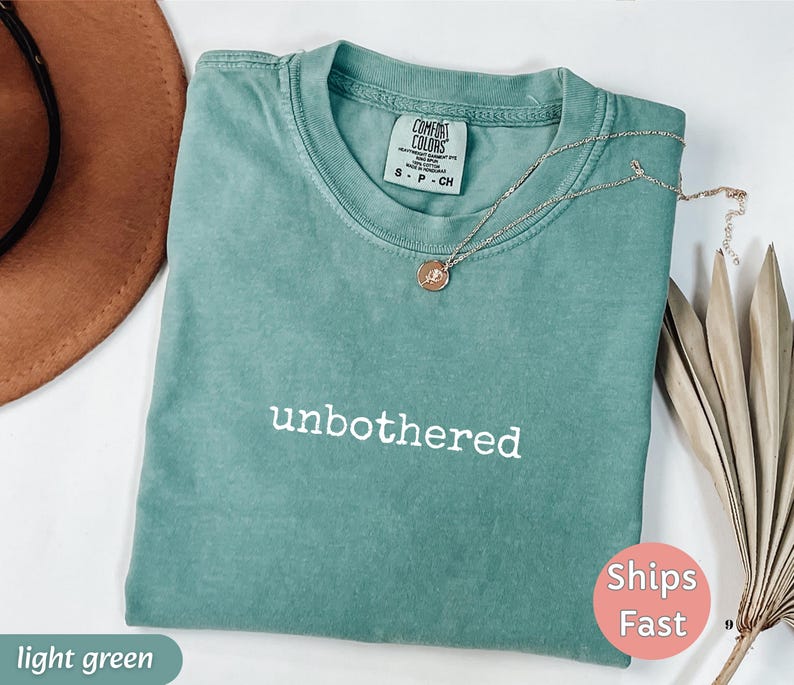 May include: A light green t-shirt with the word "unbothered" in white lettering. A gold necklace with a pendant rests on the shirt. The shirt is folded, and a brown hat is partially visible in the background.