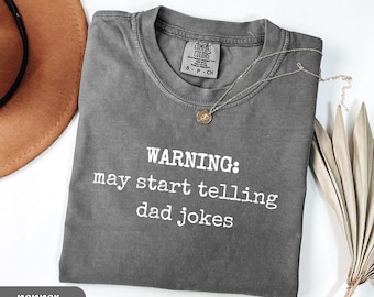 May include: Pepper-colored t-shirt with the text "WARNING: may start telling dad jokes" in white. The shirt has a crew neck and short sleeves. A gold necklace with a pendant is draped on the shirt. A brown hat is in the background.