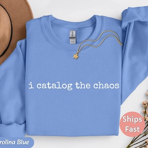 May include: A Carolina blue sweatshirt with the text "i catalog the chaos" in white. A gold necklace with a star pendant is draped on the sweatshirt. The sweatshirt has a crew neck and long sleeves.