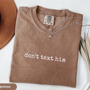 May include: Espresso-colored t-shirt with the phrase "don't text him" in white block letters. A silver necklace with a small pendant rests on the shirt. The shirt is folded, and a brown hat is visible in the background.