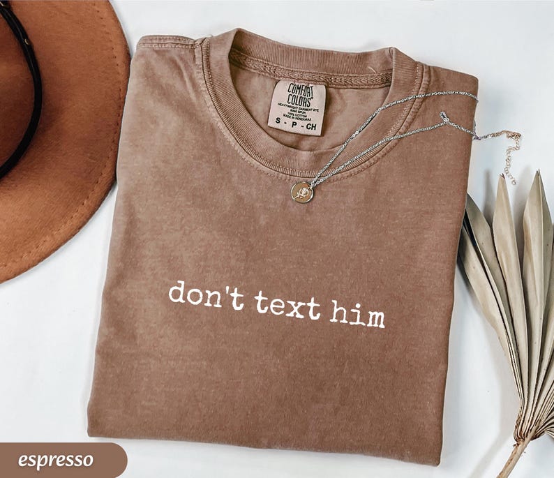 May include: Espresso-colored t-shirt with the phrase "don't text him" in white block letters. A silver necklace with a small pendant rests on the shirt. The shirt is folded, and a brown hat is visible in the background.