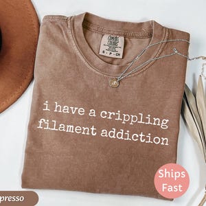 May include: Espresso-colored t-shirt with the text "i have a crippling filament addiction" in white. The shirt has a crew neck and a small tag at the neckline. A silver necklace with a pendant is draped across the shirt.