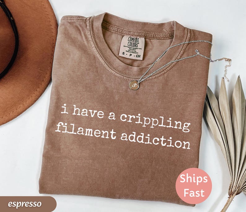 May include: Espresso-colored t-shirt with the text "i have a crippling filament addiction" in white. The shirt has a crew neck and a small tag at the neckline. A silver necklace with a pendant is draped across the shirt.