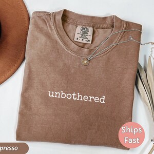 May include: Espresso-colored t-shirt with the word "unbothered" in white. The shirt has a crew neck and short sleeves. A silver necklace with a small pendant rests on the shirt. The shirt is folded and displayed with a hat and dried leaves.