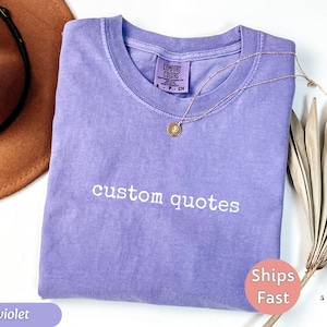 K&ouml;nnte beinhalten: Ein violettes T-Shirt mit den wei&szlig; gestickten Worten "custom quotes". Eine goldene Halskette mit Anh&auml;nger liegt auf dem Shirt. Ein brauner Hut und eine getrocknete Pflanze befinden sich im Hintergrund.